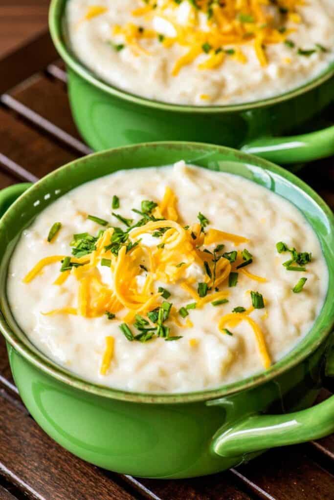 Two small creamic bowls with handles filled with potoato soup topped with chives and grated cheddar cheese.  