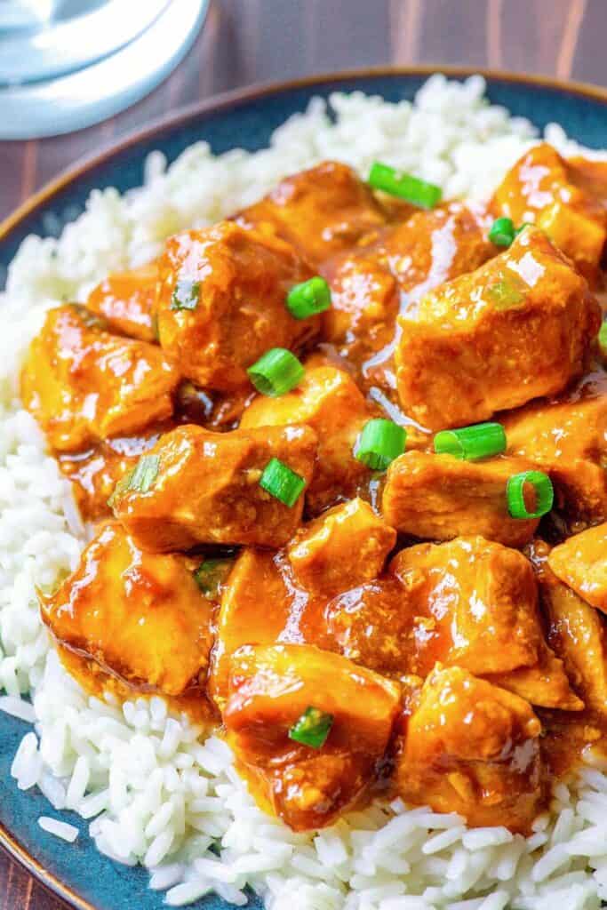 Close up of a plate of white rice topped with cubes of crockpot bourbon chicken and green onion slices. 