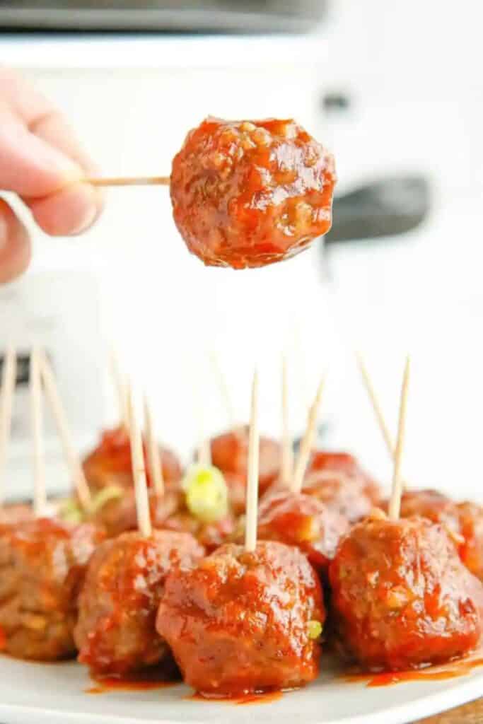 A hand uses a toothpick to pick up a slow cooker cranberry meatball coated in a glossy red cranberry sauce. More saucy meatballs with toothpicks are piled on a white serving plate below for an easy warm appetizer.