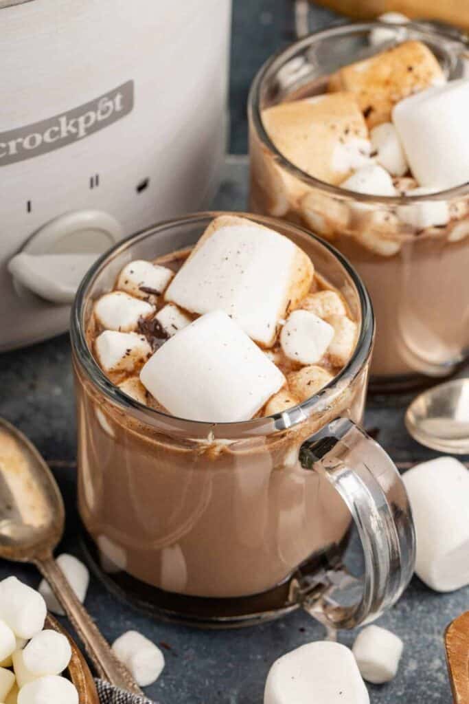 Two clear mugs filled with creamy Crockpot hot chocolate, topped with large toasted marshmallows and mini marshmallows, with a slow cooker and scattered marshmallows in the background for a cozy winter treat.