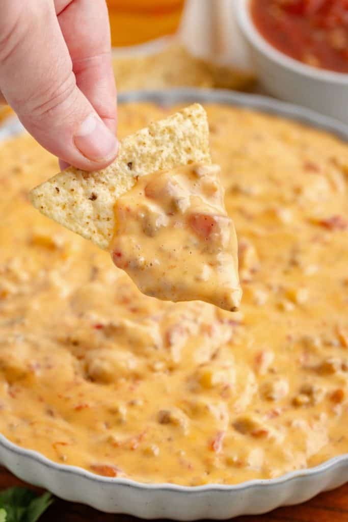 A tortilla chip scoops up creamy Crockpot Rotel Sausage Dip, loaded with melted cheese, tomatoes, and sausage. The hearty dip fills a shallow dish.