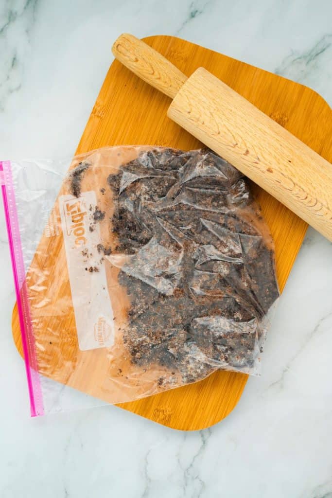 Plastic zipper bag filled with crushed chocolate sandwich cookies resting on a wooden cutting board, with a rolling pin beside it used for breaking up the cookies into crumbs.