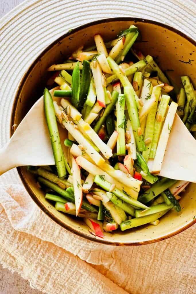 A fresh cucumber apple salad served in a rustic yellow bowl, made with thin matchstick slices of cucumber and red apple, sprinkled with sunflower seeds and fresh dill. Wooden salad tongs rest inside the bowl, placed on a textured neutral cloth for a cozy presentation.
