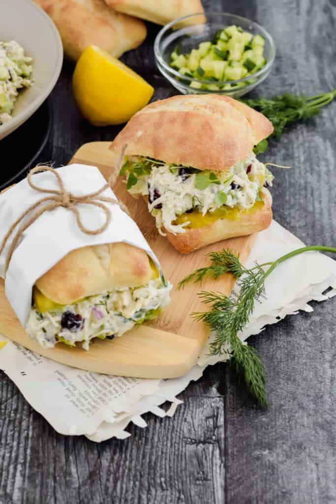 Chicken salad sandwiches served on soft ciabatta rolls, filled with a creamy chicken salad mixed with diced celery, red onion, and dried cranberries. The sandwiches are topped with fresh greens and paired with lemon and herbs.