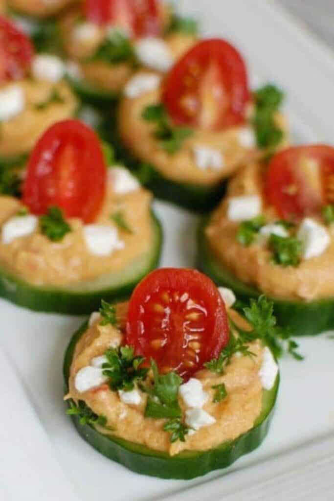 Thick cucumber slices topped with cucumber hummus bites made with a creamy spread and garnished with halved cherry tomatoes fresh herbs and small crumbles of cheese. The bite sized appetizers are arranged on a plate highlighting their fresh colorful presentation and layered textures.