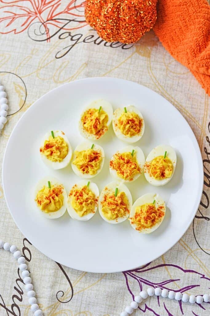 Overhead view of cute pumpkin shaped deviled eggs arranged on a white plate with creamy yellow filling dusted with smoked paprika and topped with chive stems. An orange decorative pumpkin and Fall table setting make this a festive Thanksgiving appetizer.