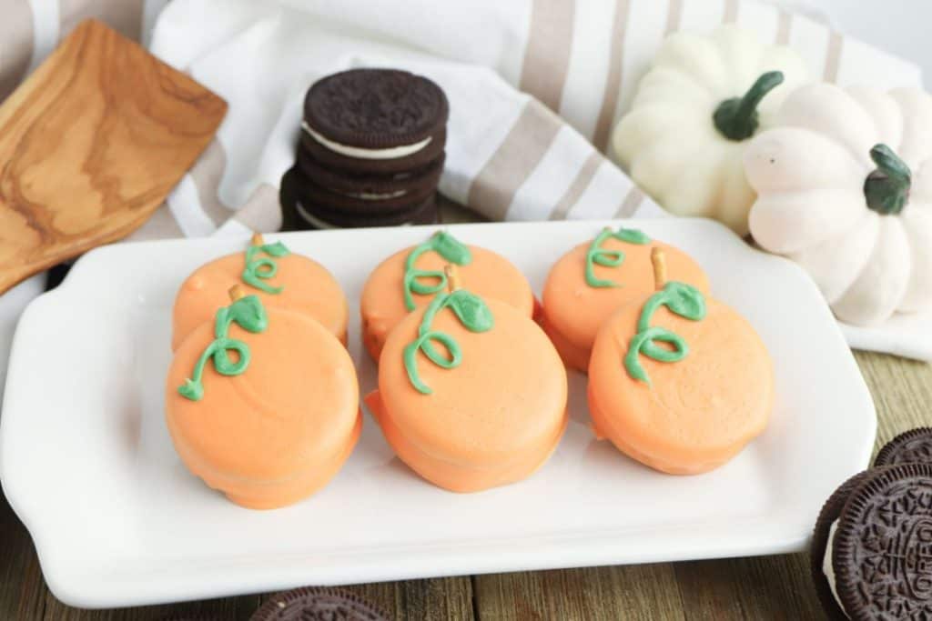 Six dipped Oreos decorated with green icing vines and stems are arranged on a white plate. The cookies are coated in orange candy to resemble pumpkins, with whole Oreos and white pumpkins in the background for a festive fall touch.