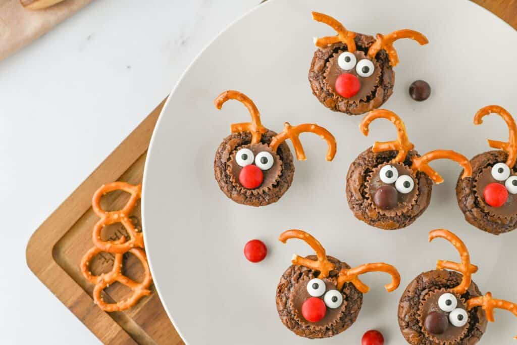 Plate of festive reindeer themed brownie bites decorated with candy eyes, red or brown candy noses, peanut butter cups, and pretzel antlers. Extra pretzels and candies are scattered on a wooden cutting board and white surface beside the plate.