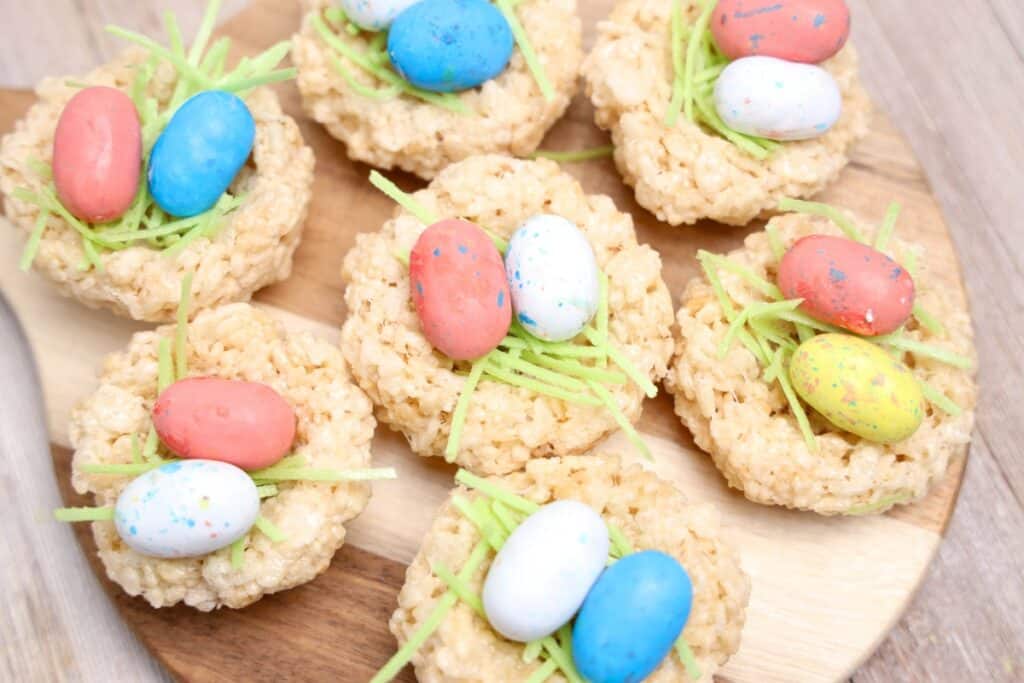 Round Easter Rice Krispie treats shaped like small nests sit on a wooden board. Each nest is topped with green candy grass and colorful speckled chocolate eggs in pastel pink blue yellow and white.