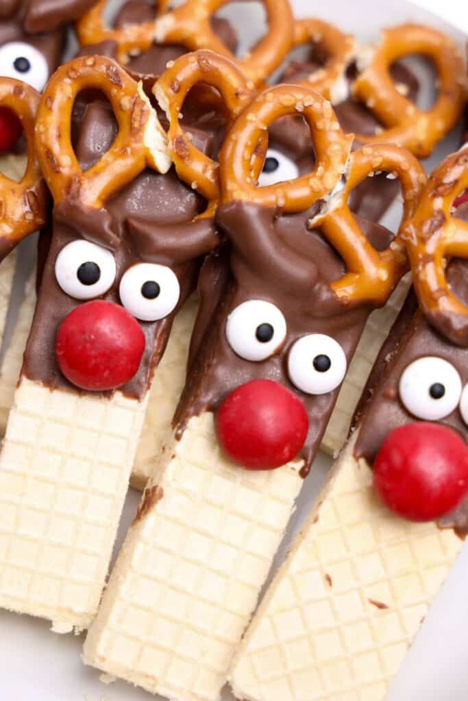 Close-up of completed cute Rudolph cookies made with vanilla wafer bars dipped in chocolate, featuring wide candy eyes, red M&M noses, and mini pretzels as antlers. The cookies are bunched together on a plate.