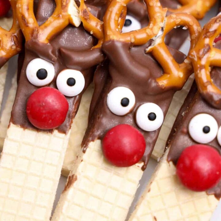 Close-up of completed cute Rudolph cookies made with vanilla wafer bars dipped in chocolate, featuring wide candy eyes, red M&M noses, and mini pretzels as antlers. The cookies are bunched together on a plate.