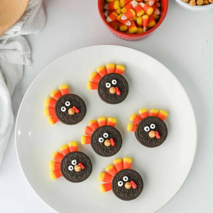 Overhead view of a white plate with six fun Thanksgiving treats for kids, featuring Oreo turkey cookies decorated with candy corn feathers, candy eyes, orange candy beaks, and red icing wattles. Surrounded by festive ingredients like candy corn, butterscotch chips, and a pumpkin for a playful fall setup.