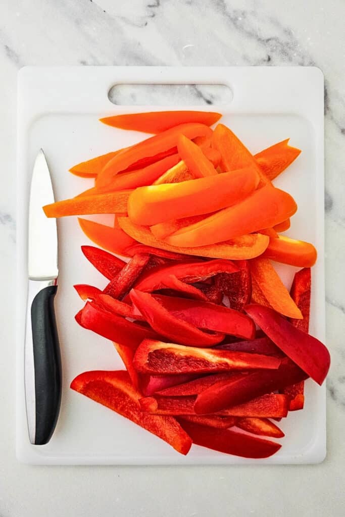 Red and orange bell peppers are sliced into long strips and arranged on a white cutting board. A kitchen knife rests beside the prepared peppers on a marble countertop.