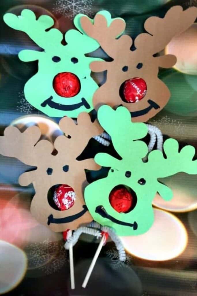 Paper cutouts shaped like reindeer heads feature a red sucker in the center as the nose, with hand-drawn eyes and smiles. These festive reindeer lollipop holders come in green and brown paper and are a fun holiday craft and treat combo.