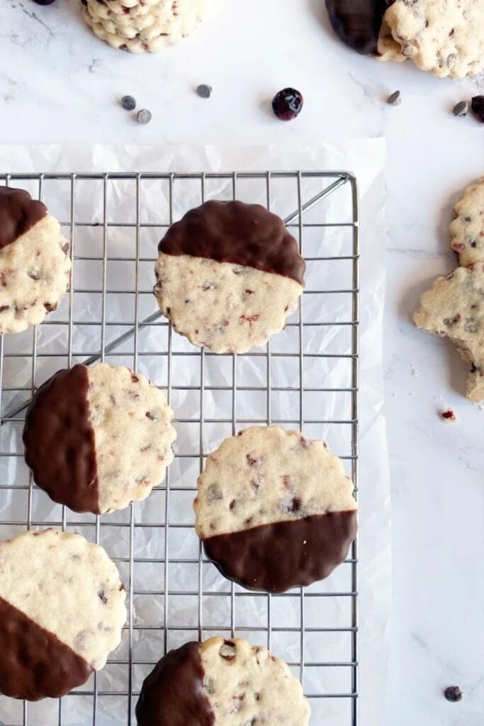 Round cranberry shortbread cookies, half-dipped in glossy dark chocolate, displayed on a cooling rack over parchment paper. The buttery cookies are speckled with bits of dried cranberry and mini chocolate chips.