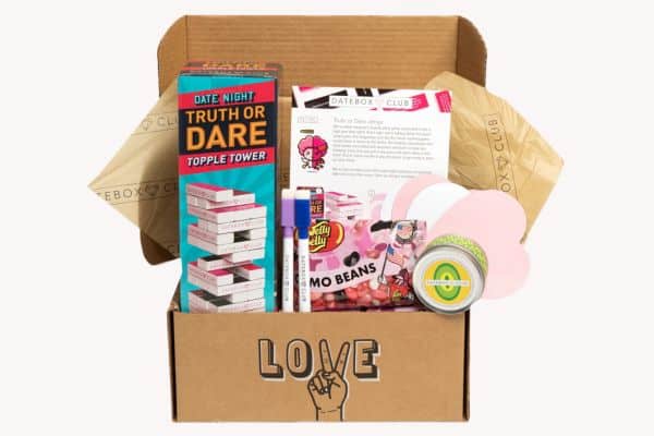 Open DateBox Club subscription box featuring a "Date Night Truth or Dare Topple Tower" game, a pack of Jelly Belly Emo Beans, a date night activity card, lip balm, markers, and pink heart-shaped paper cutouts, all packed in a box labeled “LOVE” with hand sign art. The items are curated to encourage playful and romantic connection.