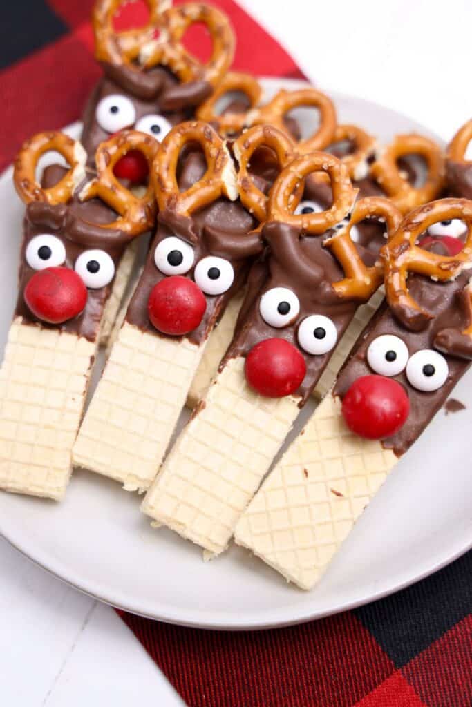 Plate of decorated reindeer cookie snacks made from vanilla wafer bars partially dipped in chocolate, with candy eyes, red M&M noses, and pretzel twists for antlers. The playful holiday treats are arranged on a white plate over a red and black plaid napkin.