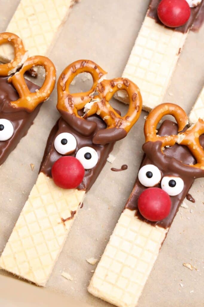 Finished reindeer cookies on parchment paper, made from vanilla wafers dipped in chocolate, decorated with candy eyes, red M&M noses, and pretzel antlers.
