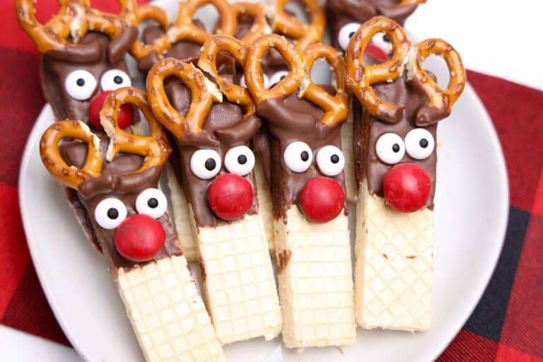 Plate of festive reindeer cookies made from vanilla wafer bars dipped in chocolate, decorated with candy eyes, red chocolate noses, and mini pretzels as antlers. The cookies are arranged closely on a white plate atop a red and black checkered cloth.