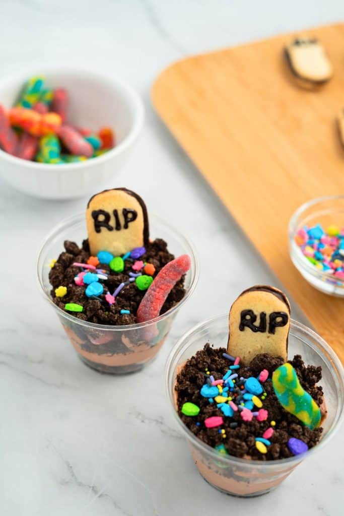 Two Halloween-themed dirt cups made with layers of chocolate pudding and crushed cookies, topped with Milano cookie tombstones labeled “RIP,” colorful spooky sprinkles, and gummy worms. A bowl of extra gummy worms and decorating supplies are visible in the background.