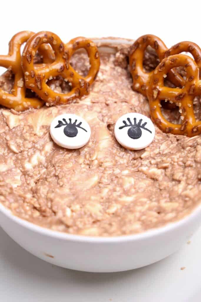 Partially decorated Christmas dip in a white bowl, featuring candy eyes with lashes and mini pretzel antlers placed on top. The nutella and cream cheese mixture is swirled and textured, ready for the final festive touches.
