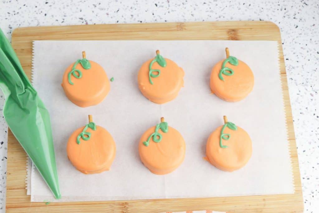 Six pumpkin-themed treats made from orange candy-coated oreo cookies are arranged on parchment paper over a wooden cutting board. Each cookie has a pretzel stick stem and green icing vine, with a green piping bag resting on the left side.
