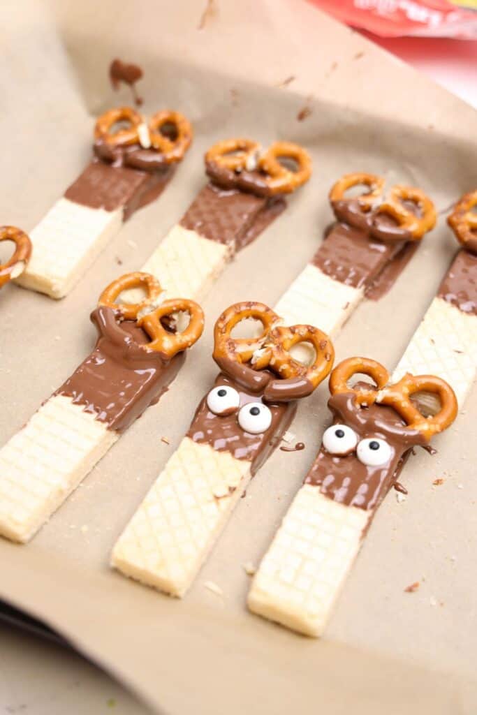 Partially decorated reindeer cookies laid out on parchment paper, featuring vanilla wafer bars dipped in chocolate with pretzel antlers and candy eyes beginning to be added. The treats are mid-assembly, showing the step-by-step process of creating the Christmas snacks.