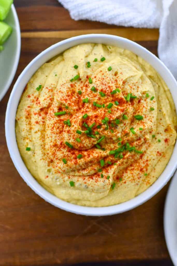 Smooth and creamy deviled egg dip in a white bowl, topped with a sprinkle of paprika and finely chopped chives. The dip has a rich yellow hue and swirled texture, giving it a classic deviled egg appearance in dip form.