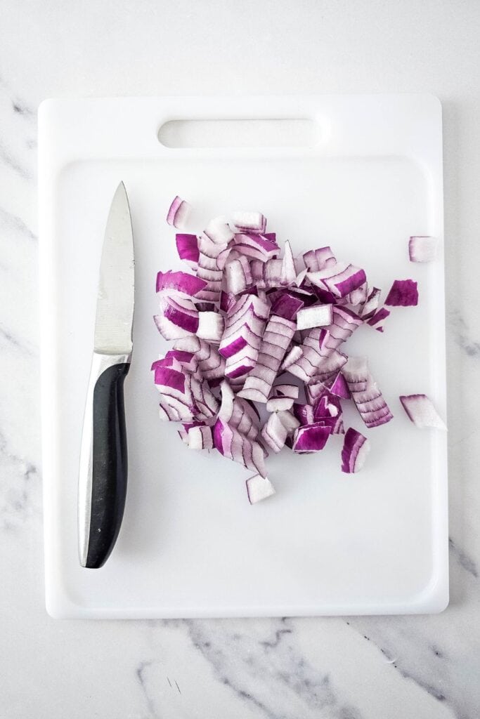Diced red onions for pasta salad on a white plastic cutting board next to a small paring knife with a black handle, all set on a light marble countertop.