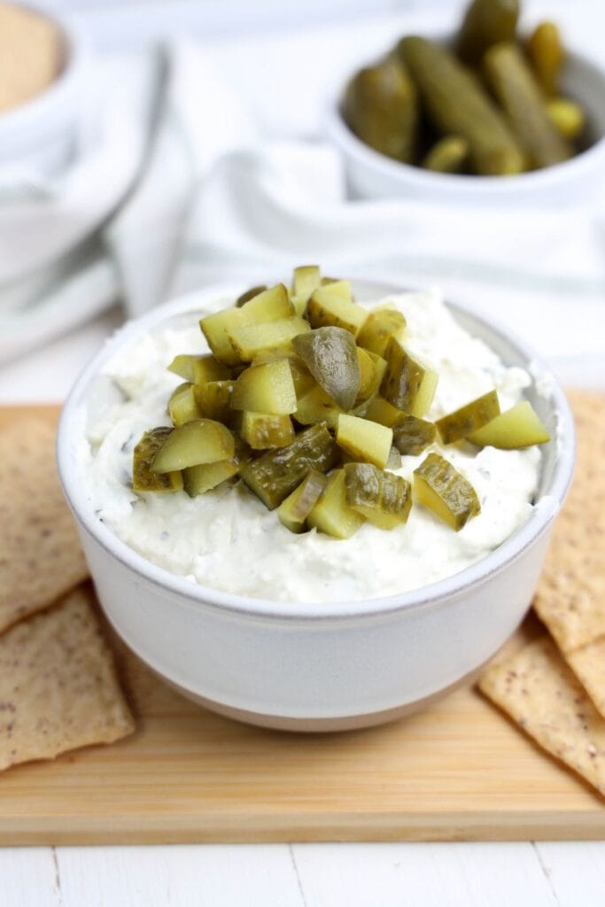 A white bowl filled with creamy dill pickle dip, generously topped with chopped pickles, sits on a wooden board surrounded by crispy crackers. A bowl of whole pickles is blurred in the background, adding a rustic and fresh touch.