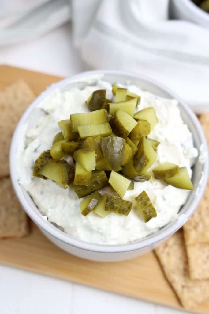 Creamy dill pickle dip topped with a generous pile of chopped dill pickles in a white bowl. Crackers are arranged around the bowl for dipping.