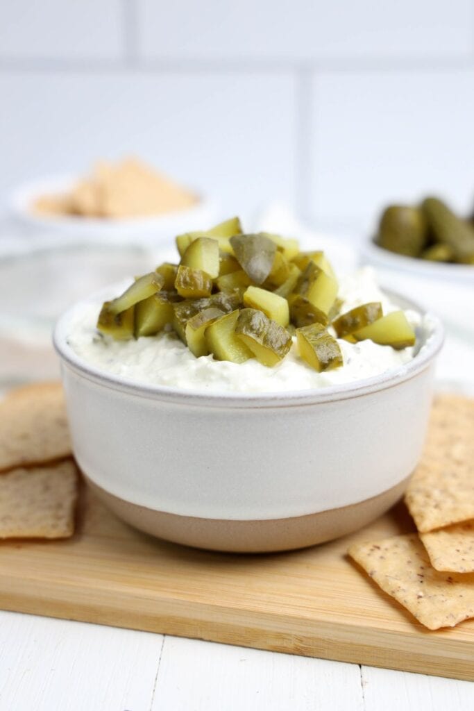 A bowl of creamy dill pickle dip topped with chopped pickles sits on a wooden board, surrounded by whole grain crackers. The dip is thick and tangy, served in a white and beige ceramic bowl.