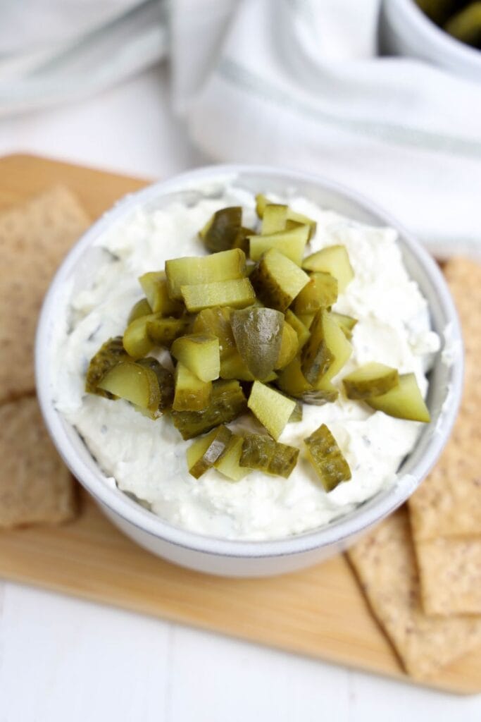 A close-up of a bowl filled with creamy, cold dill pickle dip, topped generously with chopped dill pickles. The bowl is placed on a wooden board with whole grain crackers on the side.