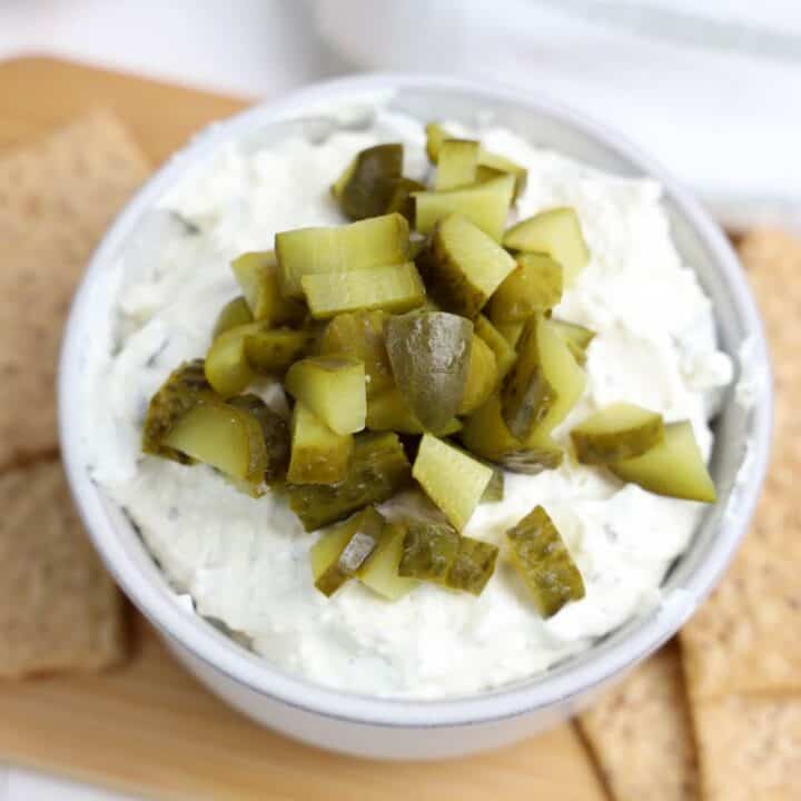 A close-up of a bowl filled with creamy, cold dill pickle dip, topped generously with chopped dill pickles. The bowl is placed on a wooden board with whole grain crackers on the side.