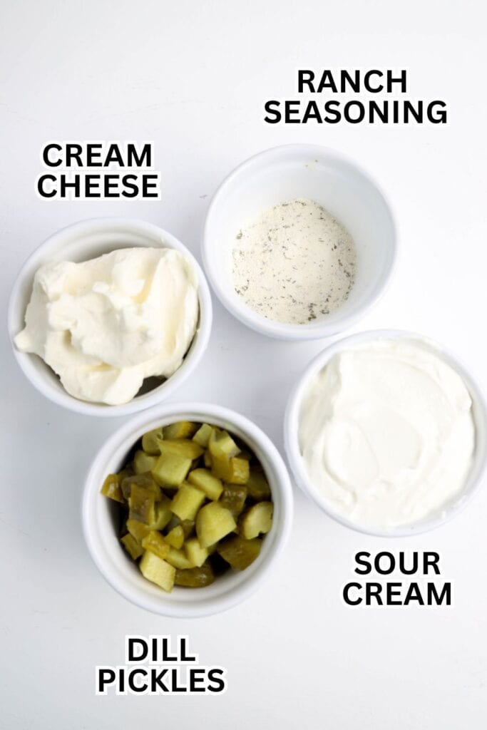 Four small white bowls arranged on a white surface, each labeled with its contents: cream cheese, ranch seasoning, sour cream, and chopped dill pickles, ingredients for a creamy dill pickle dip.