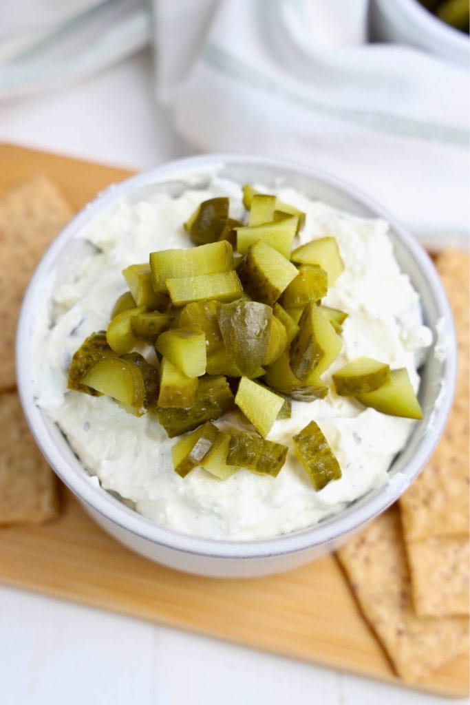 Creamy dill pickle game day party dip served in a white bowl, topped with a generous pile of chopped dill pickles. The bowl is placed on a wooden board with tortilla chips arranged nearby for dipping.