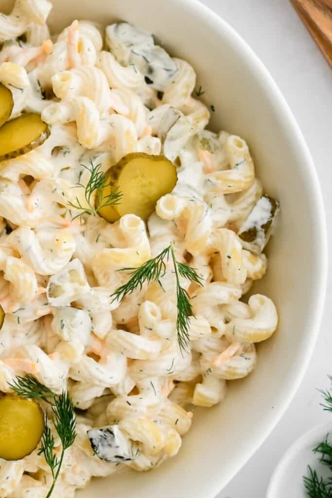 Bowl of dill pickle pasta salad with elbow macaroni chopped pickles shredded carrots and fresh dill tossed in a creamy dressing. Pickle slices and sprigs of dill on top highlight the tangy dill pickle flavor of the pasta salad.