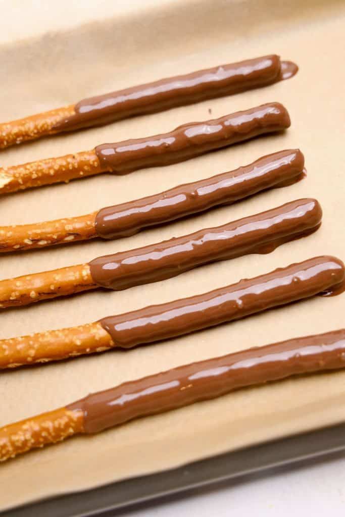 Six chocolate dipped pretzel sticks laid out on parchment paper, with the top half of each pretzel coated in smooth, glossy milk chocolate. The pretzels are arranged in neat rows.
