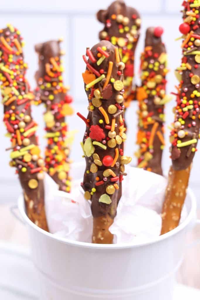 Close-up of dipped chocolate pretzels with Fall sprinkles, displayed in a white pail lined with tissue paper. Each pretzel rod is coated in chocolate and topped with festive red, orange, yellow, and gold sprinkles shaped like leaves and dots, creating a vibrant autumn treat.