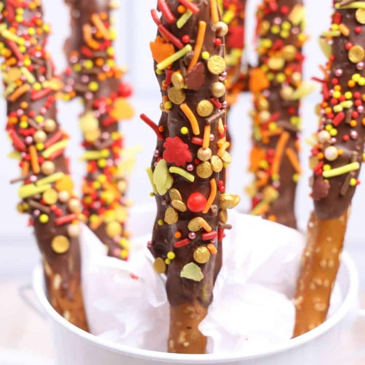 Close-up of dipped chocolate pretzels with Fall sprinkles, displayed in a white pail lined with tissue paper. Each pretzel rod is coated in chocolate and topped with festive red, orange, yellow, and gold sprinkles shaped like leaves and dots, creating a vibrant autumn treat.