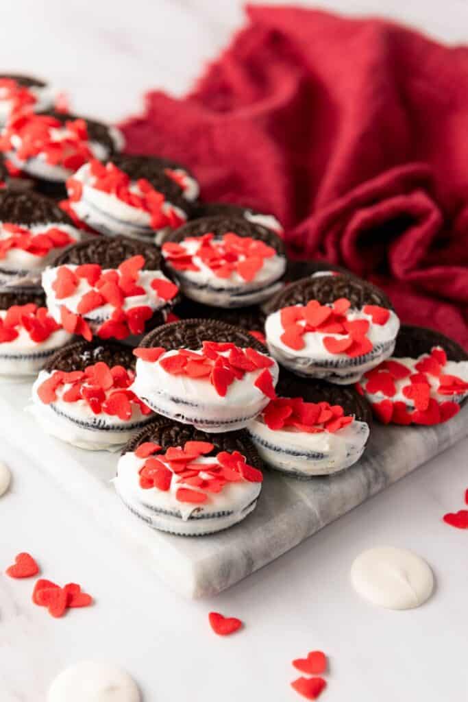 Pile of white chocolate dipped oreos with candy heart sprinkles on a marble cutting board. 