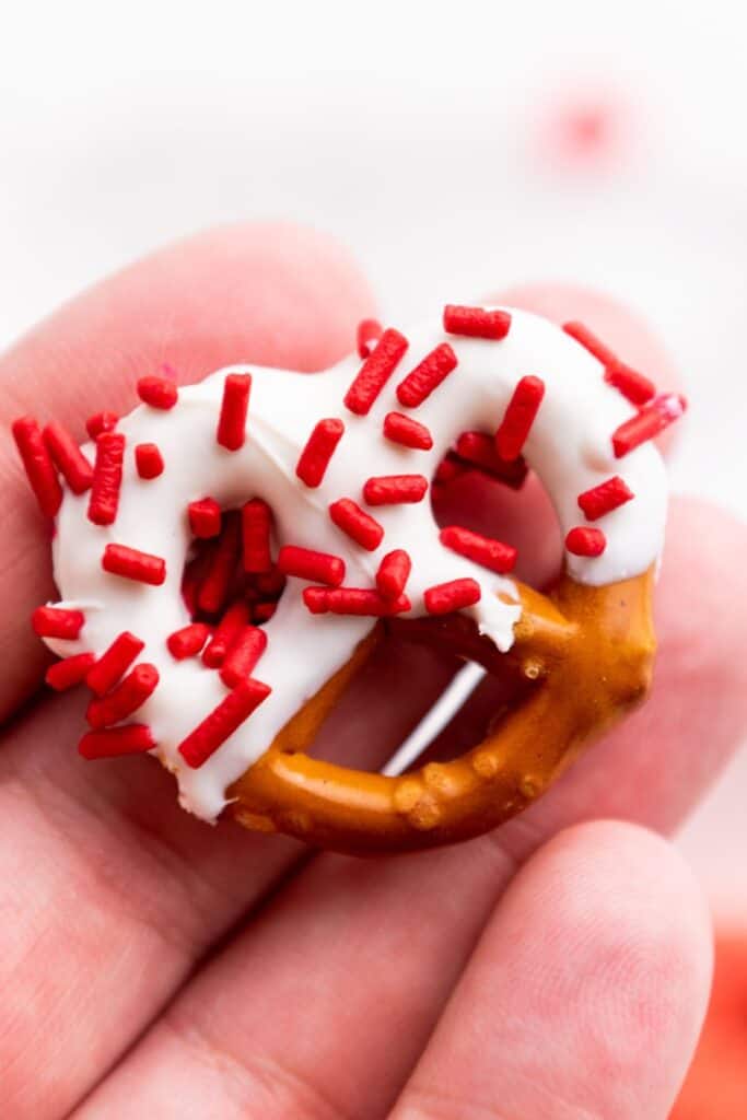 Close up of a hand holding a mini pretzel dipped in white candy melt and red sprinkles. 