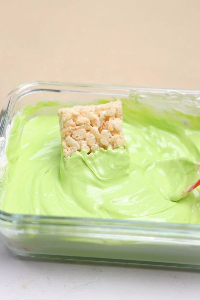 A Rice Krispie treat partially dipped into a bowl of smooth, melted green candy coating inside a glass dish. The treat is being coated for decorating Frankenstein treats. 