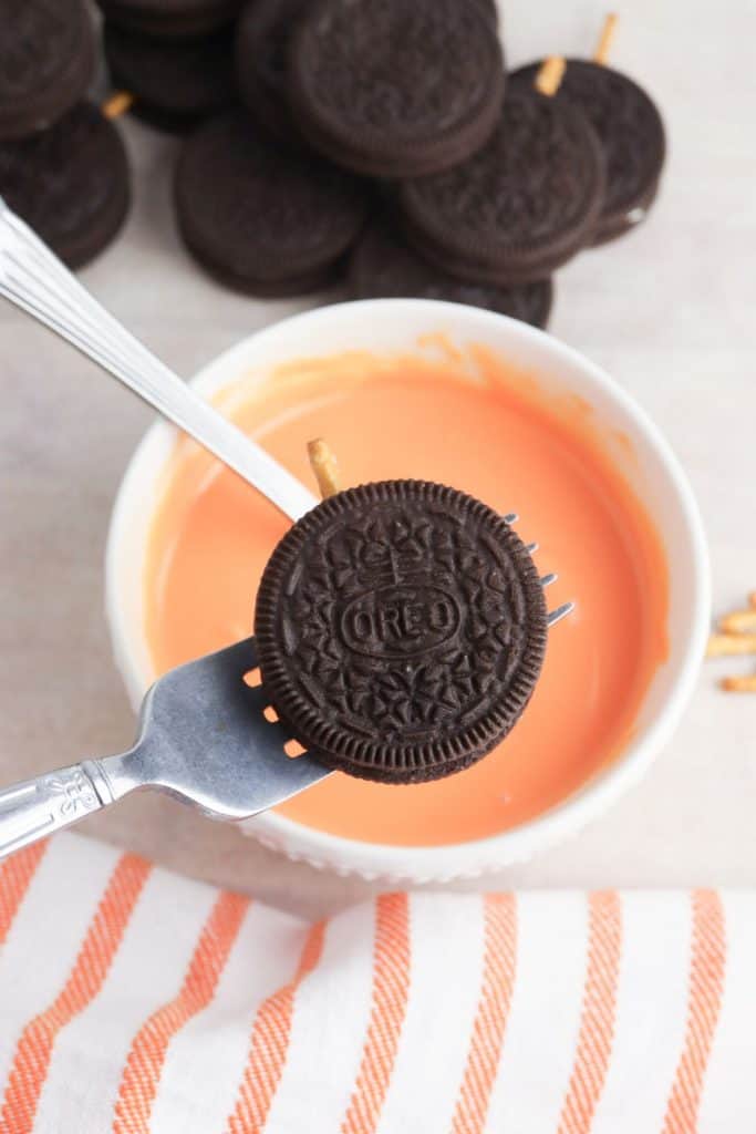 A chocolate sandwich cookie with a pretzel stick inserted is held over a bowl of melted orange candy using a fork. The setup rests on parchment paper with more cookies and a striped orange and white towel visible in the background.