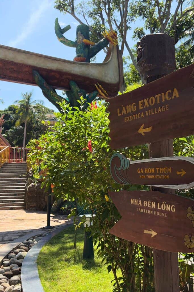 Wooden directional signs at Hon Thom Park in Phu Quoc, Vietnam, point toward Exotica Village, Hon Thom Station, and Lantern House, surrounded by tropical greenery. In the background, a whimsical sculpture of a green creature paddling a canoe arches above a pathway lined with trees and stone steps.