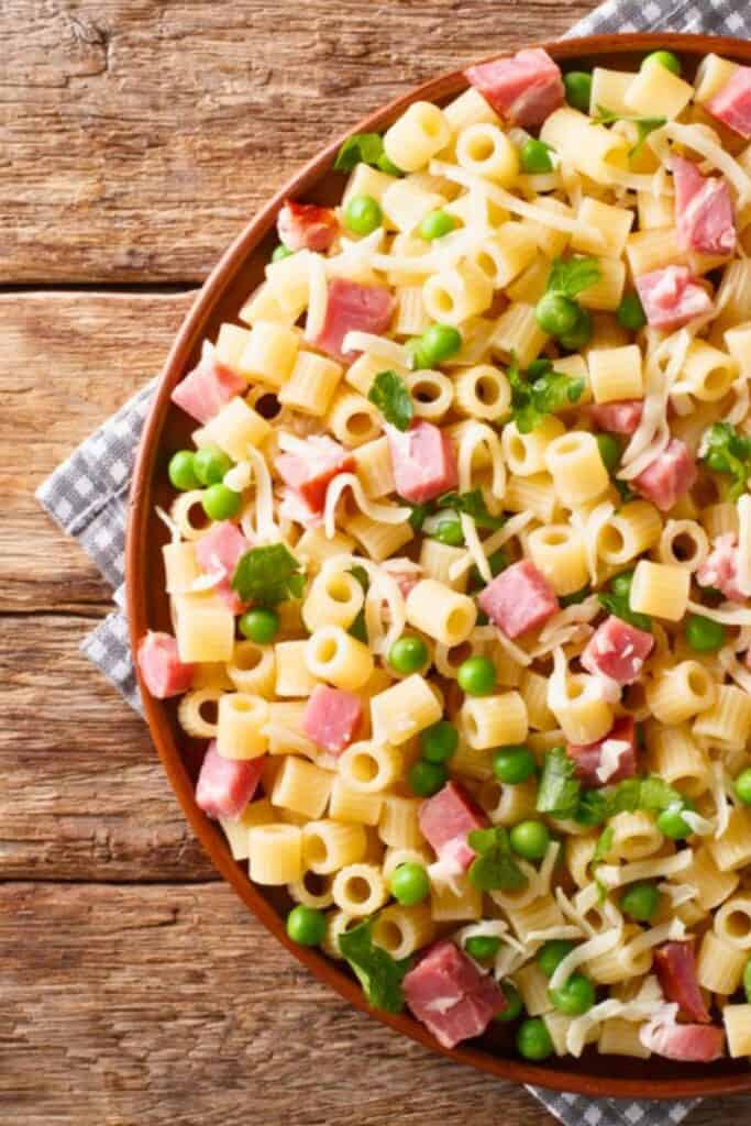 Bowl of ditalini pasta salad with small tube pasta green peas diced ham shredded cheese and parsley served on a rustic wooden table. The simple cold pasta salad highlights classic deli style ingredients mixed with tender pasta.