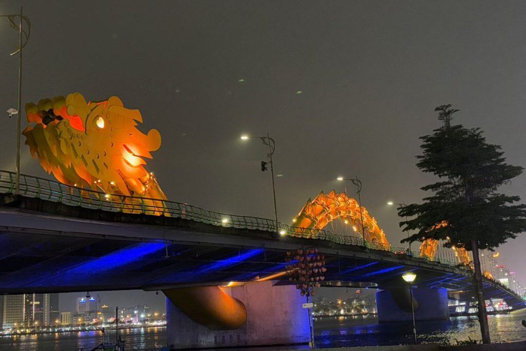 The Dragon Bridge in Da Nang is illuminated at night, with its massive golden dragon structure glowing brightly against the dark sky. The bridge stretches over the Han River, with reflections of city lights shimmering in the water below.