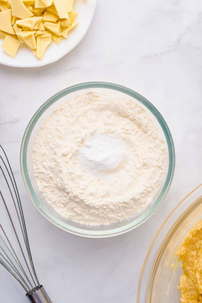 Glass bowl filled with all-purpose flour and baking soda, ready to be mixed into cookie dough, with chopped white chocolate nearby and part of a mixing bowl and whisk visible on the side.