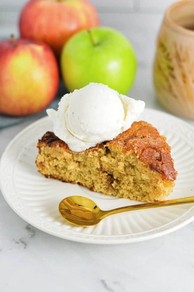 A slice of Dutch apple cake served on a white plate, topped with a scoop of vanilla ice cream. The golden, slightly crisp exterior and moist, spiced crumb pair perfectly with the fresh apples in the background.