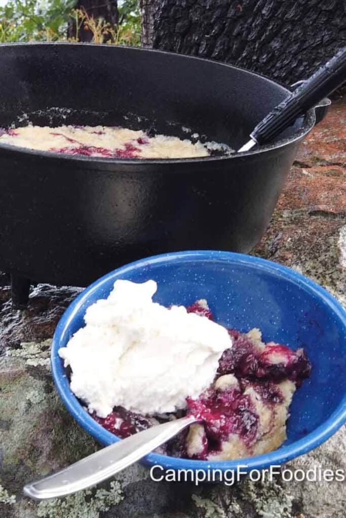Blue bowl filled with warm campfire berry cobbler topped with a scoop of whipped cream, served outdoors beside a cast iron pot. The dessert shows bubbling purple berries and soft biscuit topping, making it a cozy camping treat.